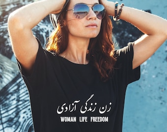 以下が含まれることがあります： 白い文字で「زن زندگی آزادی」と書かれた黒いTシャツを着ている人。英語では「Woman Life Freedom」と訳されます。その人はサングラスをかけていて、両手で頭を支えています。