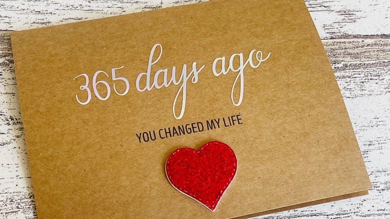 May include: A brown card with the text "365 days ago you changed my life" in white lettering. A red glitter heart is attached to the card.