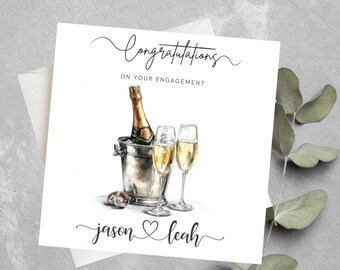 May include: Engagement card with watercolor illustration. Features a champagne bottle in a silver bucket, two champagne flutes, and wedding rings. The card reads "Congratulations on Your Engagement" and includes the names "Jason & Leah".