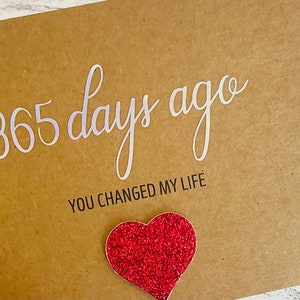 May include: A brown card with the text "365 days ago you changed my life" in white lettering. A red glitter heart is attached to the card.