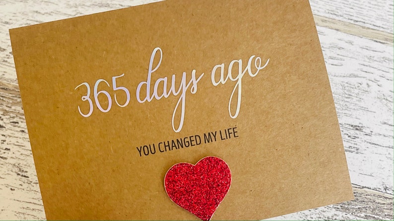 May include: A brown card with the text "365 days ago you changed my life" in white lettering. A red glitter heart is attached to the card.