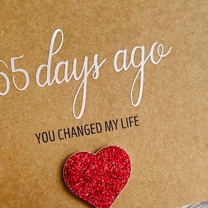 May include: A brown card with the text "365 days ago you changed my life" in white and black lettering. A red glitter heart is attached to the bottom of the card.