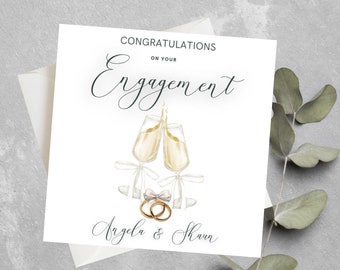 May include: A white card with the text "Congratulations on your Engagement" in black script. Two champagne flutes clink together with a white bow and two gold rings below. The names "Angela & Shawn" are written in black script below the rings.