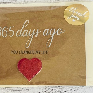 May include: A brown card with the text "365 days ago you changed my life" and a red glitter heart. A gold sticker with the text "thank you" is on the top right corner of the card.