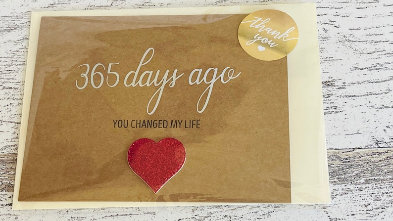 May include: A brown card with the text "365 days ago you changed my life" and a red glitter heart. A gold sticker with the text "thank you" is on the top right corner of the card.