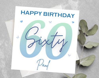 Può includere: Un biglietto bianco con un numero "60" sfumato in blu e verde con la parola "Sessanta" scritta in corsivo blu. Il testo "Buon compleanno" è scritto in blu nella parte superiore del biglietto. Il nome "Paul" è scritto in corsivo blu nella parte inferiore del biglietto.