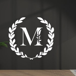 May include: A white letter "M" is centered on a dark grey wall, encircled by a laurel wreath design. A potted palm tree sits to the left. The room has a large window with blinds, allowing natural light to enter.