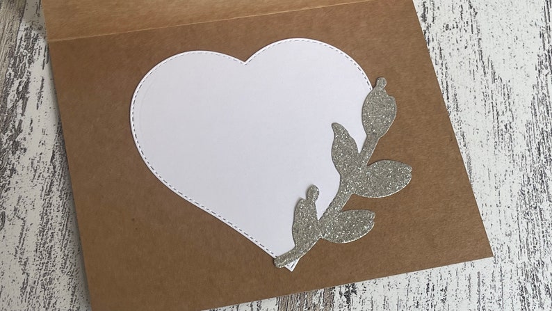 May include: A brown card with a white heart-shaped cutout and a silver glitter branch with leaves on the right side.