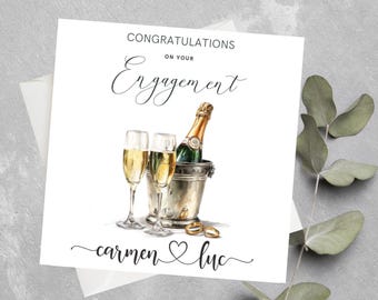 May include: White engagement card with the words "Congratulations on your Engagement". The card features a watercolor illustration of champagne glasses, a bottle of champagne in a bucket, and two rings. The names "Carmen & Luc" are written at the bottom.
