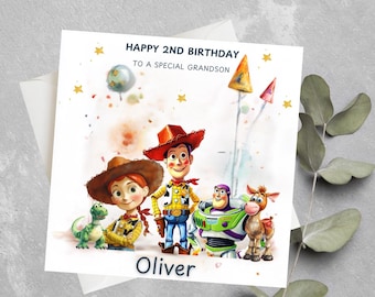 May include: A birthday card featuring a watercolor illustration of Woody, Jessie, Buzz Lightyear, and Bullseye from the Toy Story franchise. The card has a white background with gold stars and confetti. The text "Happy 2nd Birthday to a special grandson" is printed on the card. The name "Oliver" is printed at the bottom of the card.