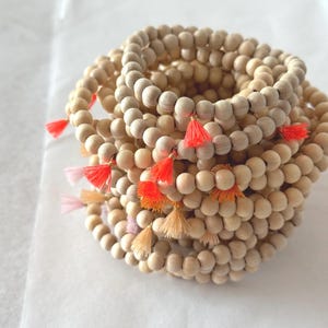 Könnte beinhalten: Ein Stapel aus mehreren kreisförmigen Armbändern aus natürlichen, hellbraunen Holzperlen. Jedes Armband ist mit einer kleinen, hängenden Quaste in verschiedenen Nuancen von Neonorange, leuchtendem Orange, zartem Pfirsich und Blassrosa versehen.