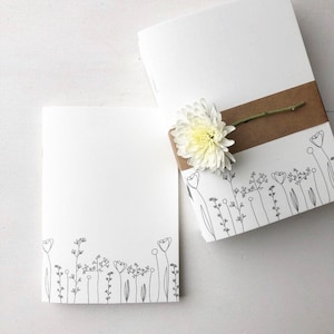 Könnte beinhalten: Zwei leere weiße Notizbücher mit einer einfachen Linienzeichnung von Blumen und Stielen am unteren Rand jeder Seite. Das Notizbuch rechts hat ein braunes Band, das um es gebunden ist, und eine weiße Blume auf dem Band.