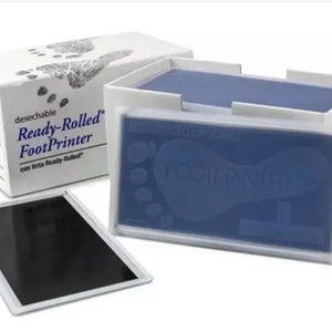 May include: A white box with the text "Ready-Rolled FootPrinter" and "desechable" on it. The box contains a blue ink pad with the text "FootPrinter" on it. There is also a black ink pad.