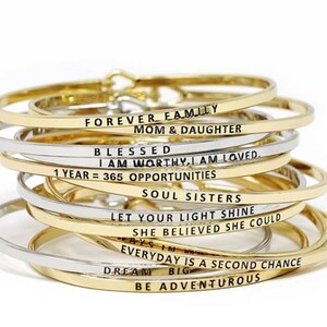 May include: A stack of gold and silver bangle bracelets with inspirational messages engraved on them. The messages include "Forever Family", "Mom & Daughter", "Blessed", "I Am Worthy, I Am Loved", "1 Year = 365 Opportunities", "Soul Sisters", "Let Your Light Shine", "She Believed She Could", "Everyday Is A Second Chance", "Dream Big", and "Be Adventurous".