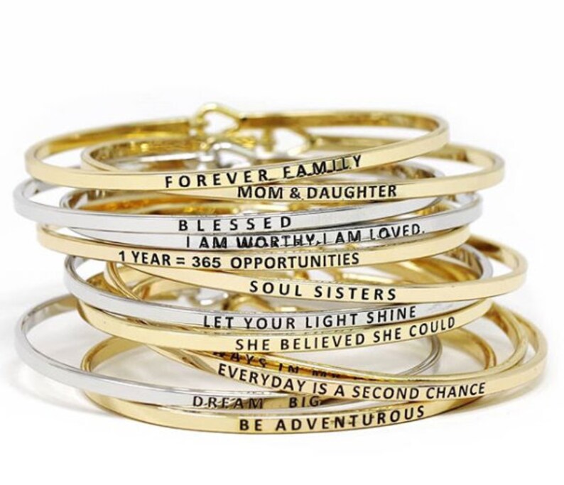 May include: A stack of gold and silver bangle bracelets with inspirational messages engraved on them. The messages include "Forever Family", "Mom & Daughter", "Blessed", "I Am Worthy, I Am Loved", "1 Year = 365 Opportunities", "Soul Sisters", "Let Your Light Shine", "She Believed She Could", "Everyday Is A Second Chance", "Dream Big", and "Be Adventurous".