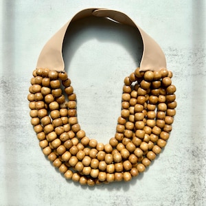 May include: A statement necklace featuring multiple strands of light brown wooden beads. The beads are strung together and attached to a wide, beige leather band that sits around the neck. The necklace is a bold, natural accessory.