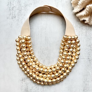 May include: A statement necklace featuring a cream-colored leather strap and a cascade of gold-colored beads. The beads are arranged in multiple rows, creating a bold and textured design. The necklace has a simple clasp closure.