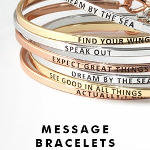 May include: A stack of gold, silver, and rose gold bangle bracelets with inspirational messages engraved on them. The messages include "Dream by the Sea", "Find Your Wing", "Speak Out", "Expect Great Things", "Dream by the Sea", "See Good in All Things", and "Actually". The bracelets are stacked on top of each other and the messages are visible on each bracelet. The bracelets are called "Message Bracelets".