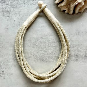 Puede incluir: Collar color crema con un diseño de cuerda trenzada. El collar está hecho de múltiples hebras de cuerda, creando un efecto en capas. Los extremos están envueltos con un material color crema a juego, y una pequeña cuenta redonda está en la parte superior.