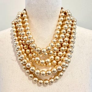 May include: A multi-strand gold-tone beaded necklace. The necklace features several rows of large, round beads in a matte gold finish. The beads are evenly spaced and create a full, layered look. The necklace is displayed on a mannequin.