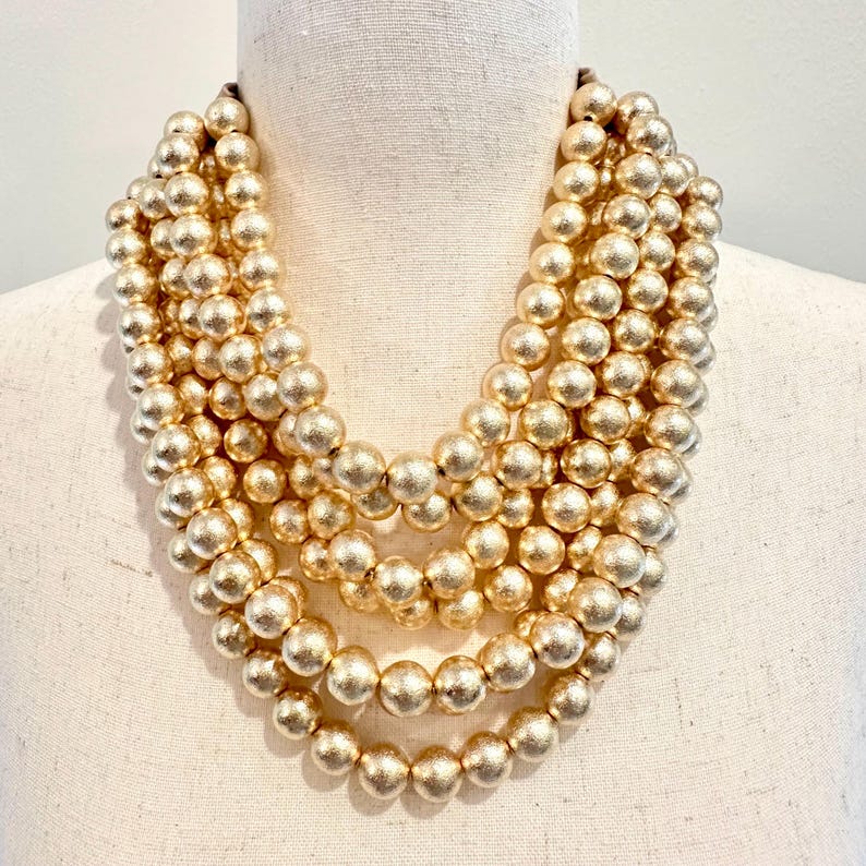 May include: A multi-strand gold-tone beaded necklace. The necklace features several rows of large, round beads in a matte gold finish. The beads are evenly spaced and create a full, layered look. The necklace is displayed on a mannequin.