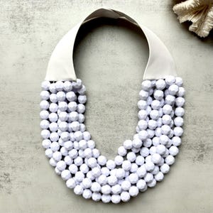 May include: A white statement necklace featuring multiple strands of faceted, round beads. The necklace has a wide, white fabric collar. The beads are a bright white color, and the necklace is displayed on a neutral background.