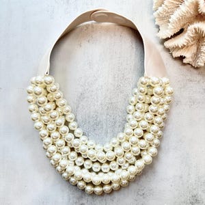 Op de afbeelding: Een witte nepparelketting met een witte elastische sluiting. De ketting is gemaakt van meerdere strengen parels en is een statement piece.