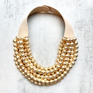 May include: A statement necklace featuring multiple strands of gold-colored beads. The beads cascade from a cream-colored leather strap that fastens with a small clasp. The necklace has a bold, eye-catching design.