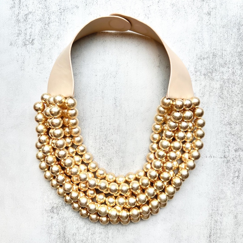 May include: A statement necklace featuring multiple strands of gold-colored beads. The beads cascade from a cream-colored leather strap that fastens with a small clasp. The necklace has a bold, eye-catching design.