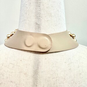 May include: A beige leather collar necklace with gold-colored spherical studs. The collar has a curved design and fastens with a matching leather closure. The necklace is displayed on a mannequin.