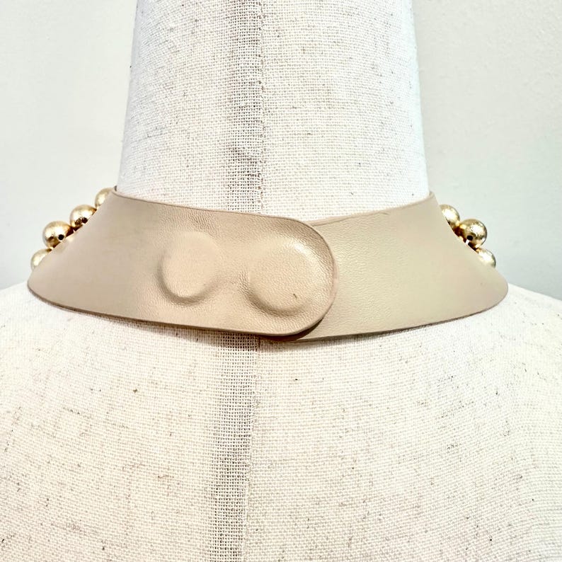 May include: A beige leather collar necklace with gold-colored spherical studs. The collar has a curved design and fastens with a matching leather closure. The necklace is displayed on a mannequin.