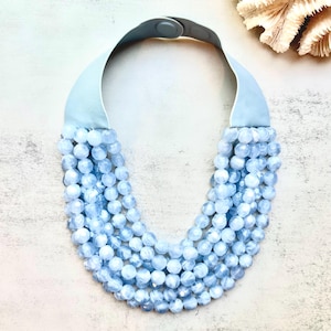 May include: A light blue beaded necklace with a pale blue leather strap and clasp. The necklace features multiple strands of faceted, round beads. The background is a light, neutral colour, with a seashell in the upper right corner.