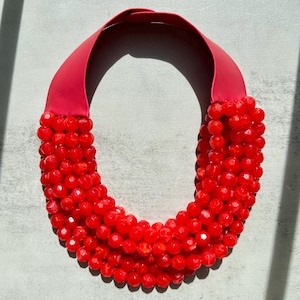 Może przedstawiać: Wyrazisty czerwony naszyjnik. Posiada szeroką, gładką czerwoną opaskę na szyję, przechodzącą w kaskadę fasetowanych czerwonych koralików. Koraliki są gęsto upakowane, tworząc odważny design. Naszyjnik jest efektownym dodatkiem.