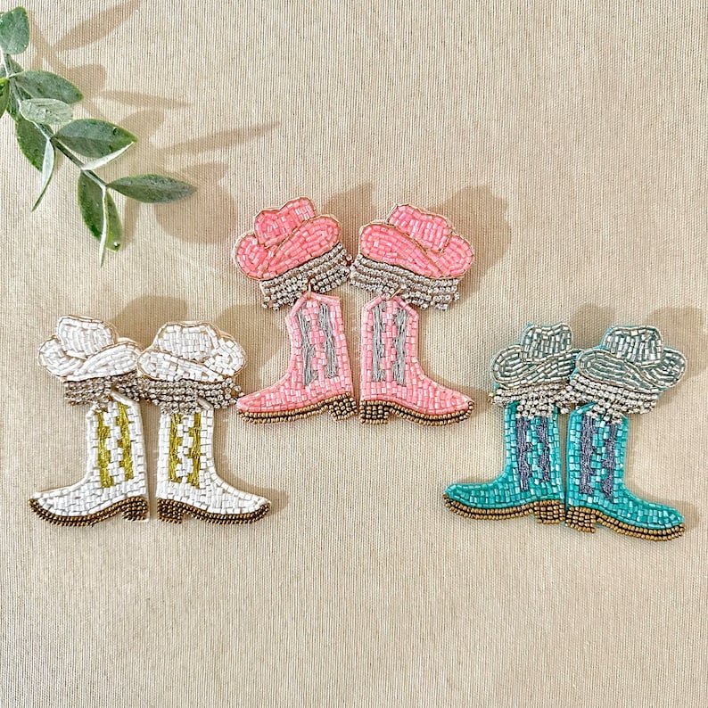 K&ouml;nnte beinhalten: Drei Paar Perlenohrringe mit Cowboy-Stiefel-Designs. Das erste Paar ist pink mit wei&szlig;en Akzenten, das zweite Paar ist wei&szlig; mit goldenen Akzenten und das dritte Paar ist t&uuml;rkis mit silbernen Akzenten.