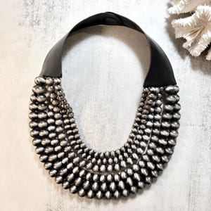 May include: A statement necklace featuring multiple strands of silver-toned beads cascading from a wide black leather collar. The beads vary in size, creating a layered effect. The necklace is designed to be worn around the neck.