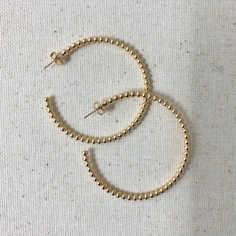 May include: A pair of gold hoop earrings made of small, round beads. The earrings are large and have a simple, elegant design.
