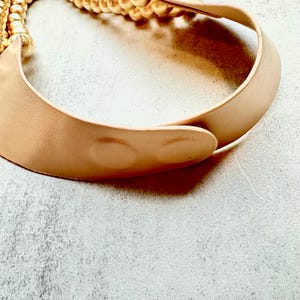 May include: A tan leather collar necklace with a gold chain detail. The necklace has a curved, crescent shape and a smooth texture. The gold chain is braided and attached to the leather collar. The necklace is shown on a light gray surface.
