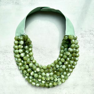 Puede incluir: Un collar de cuentas verdes con un cuello ancho de tela verde claro. El collar presenta múltiples hilos de cuentas redondas y facetadas en varios tonos de verde. El collar tiene un cierre. El collar es una pieza llamativa.