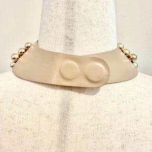 May include: A beige collar necklace with gold-colored spherical embellishments. The necklace has a curved shape and fastens with two circular buttons. The necklace is displayed on a mannequin.