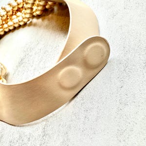 May include: A beige belt with a gold beaded accent. The belt has a smooth, leather-like texture and a curved design. The clasp features two circular, raised details. The overall aesthetic is minimalist and elegant.
