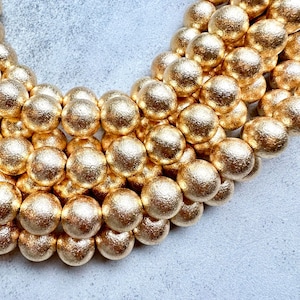 May include: Close-up of a collection of gold-colored, textured beads. The beads are spherical and arranged in a semi-circular pattern, creating a luxurious and decorative appearance. The beads are likely used for jewelry making or other craft projects.