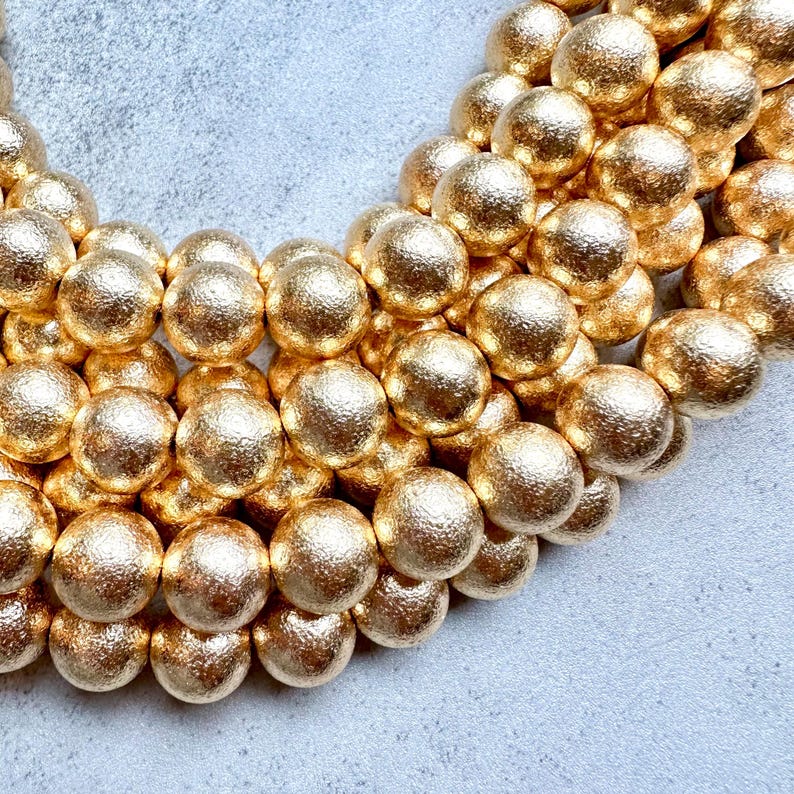 May include: Close-up of a collection of gold-colored, textured beads. The beads are spherical and arranged in a semi-circular pattern, creating a luxurious and decorative appearance. The beads are likely used for jewelry making or other craft projects.