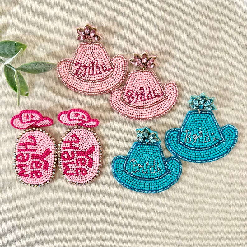 K&ouml;nnte beinhalten: Ein Set mit sechs Perlenohrringen im Cowboyhut-Design. Zwei Paare sind rosa mit dem Wort "Bride" in wei&szlig;en Perlen, und zwei Paare sind t&uuml;rkis mit dem Wort "Bride" in wei&szlig;en Perlen. Das restliche Paar ist rosa und hat ein ovales Perlenmuster.
