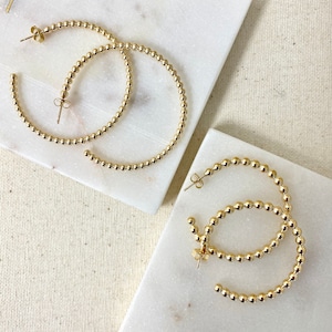 May include: A pair of gold hoop earrings made of small beads. The earrings are on a white marble surface.