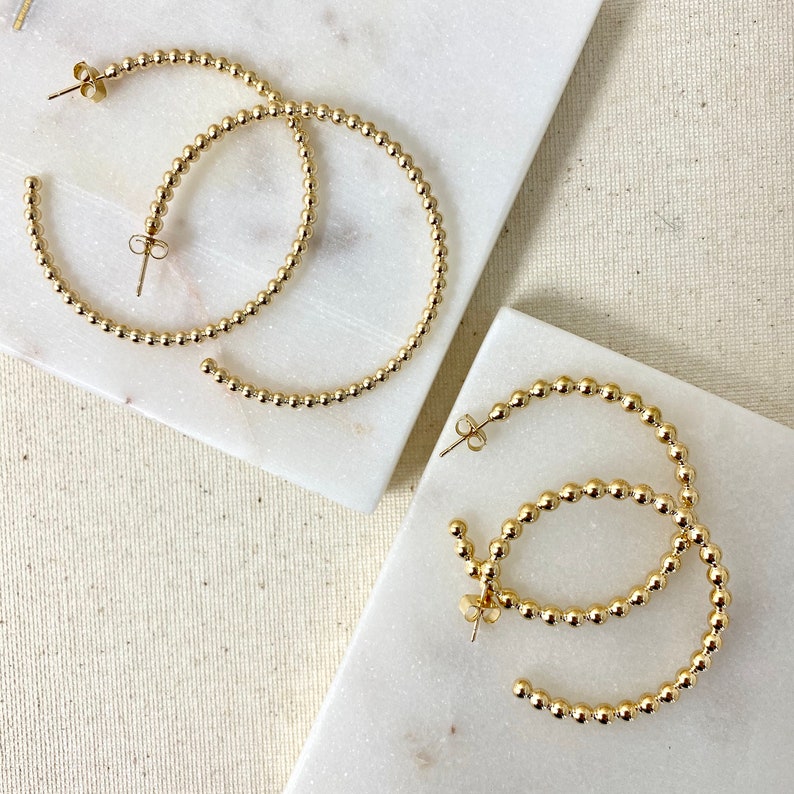 May include: A pair of gold hoop earrings made of small beads. The earrings are on a white marble surface.
