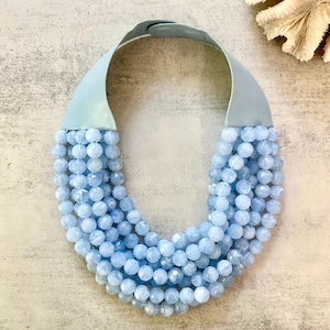 May include: A striking necklace with multiple strands of light blue faceted beads. The necklace has a wide, pale blue band that sits around the neck. The beads are a soft, pastel blue colour, creating a cascading effect.