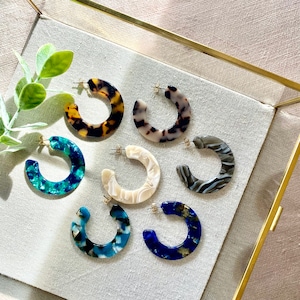 Op de afbeelding: Een set van zes kleurrijke hoepeloorbellen gemaakt van hars. De oorbellen zijn in verschillende patronen, waaronder schildpad, blauw, wit en zwart.