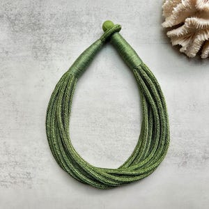 May include: A multi-strand necklace constructed from olive green cord, featuring several parallel strands that gather at the top into a single, wrapped cylindrical closure topped with a small, matching green spherical bead accent.