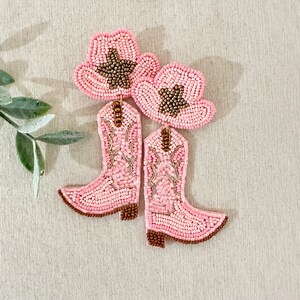 K&ouml;nnte beinhalten: Ein Paar rosa Perlenohrringe in Form von Cowboystiefeln mit einem rosa und braunen Perlen-Cowboyhut auf jedem Stiefel.