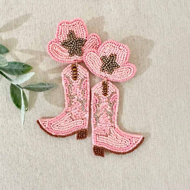 K&ouml;nnte beinhalten: Ein Paar rosa Perlenohrringe in Form von Cowboystiefeln mit einem rosa und braunen Perlen-Cowboyhut auf jedem Stiefel.